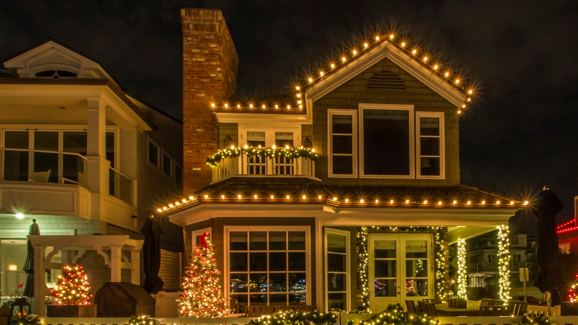 A two-story home at night outlined in warm white lights along every roofline and porch rail, with small lit trees and glowing shrubs in the front yard.